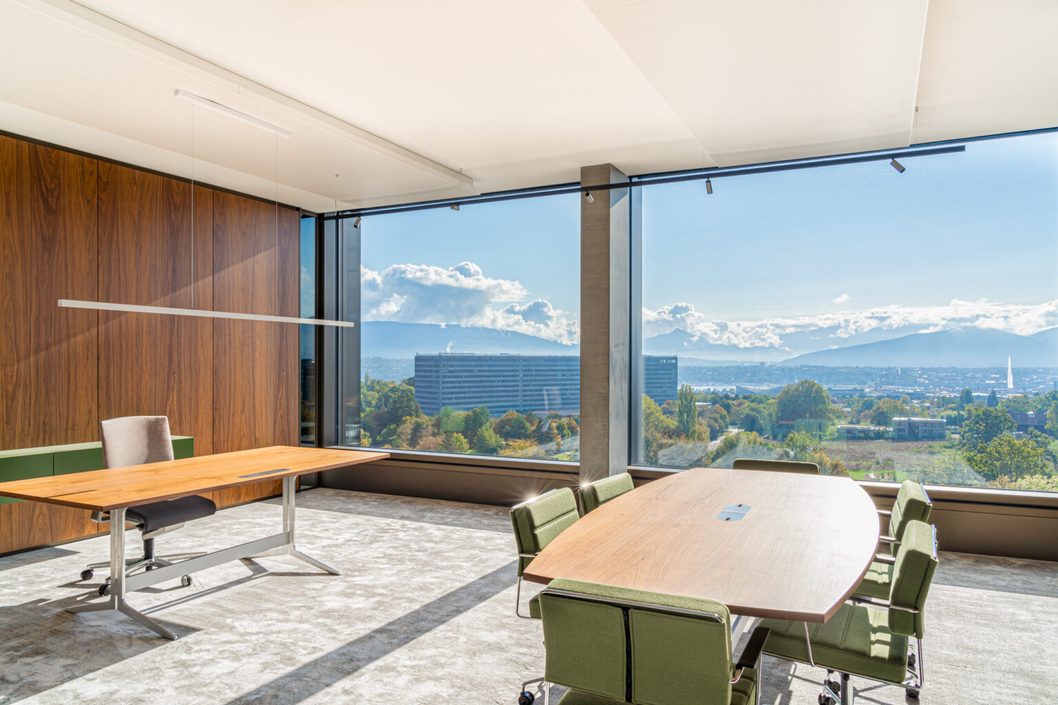 Espace de travail moderne avec bureau en bois, fauteuils verts et vue sur le bassin genevois.