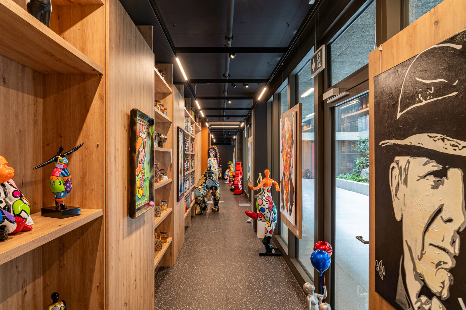 Galerie de passage bordée de baies vitrées et d'étagères exposant des œuvres pop art, peintures et sculptures colorées.