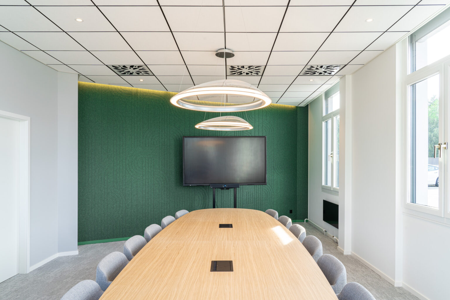 Vue frontale d'une salle de conférence baignée de lumière, dotée d'une grande table en bois, de fauteuils confortables gris, d'un écran mural et d'un mur acoustique vert foncé texturé.