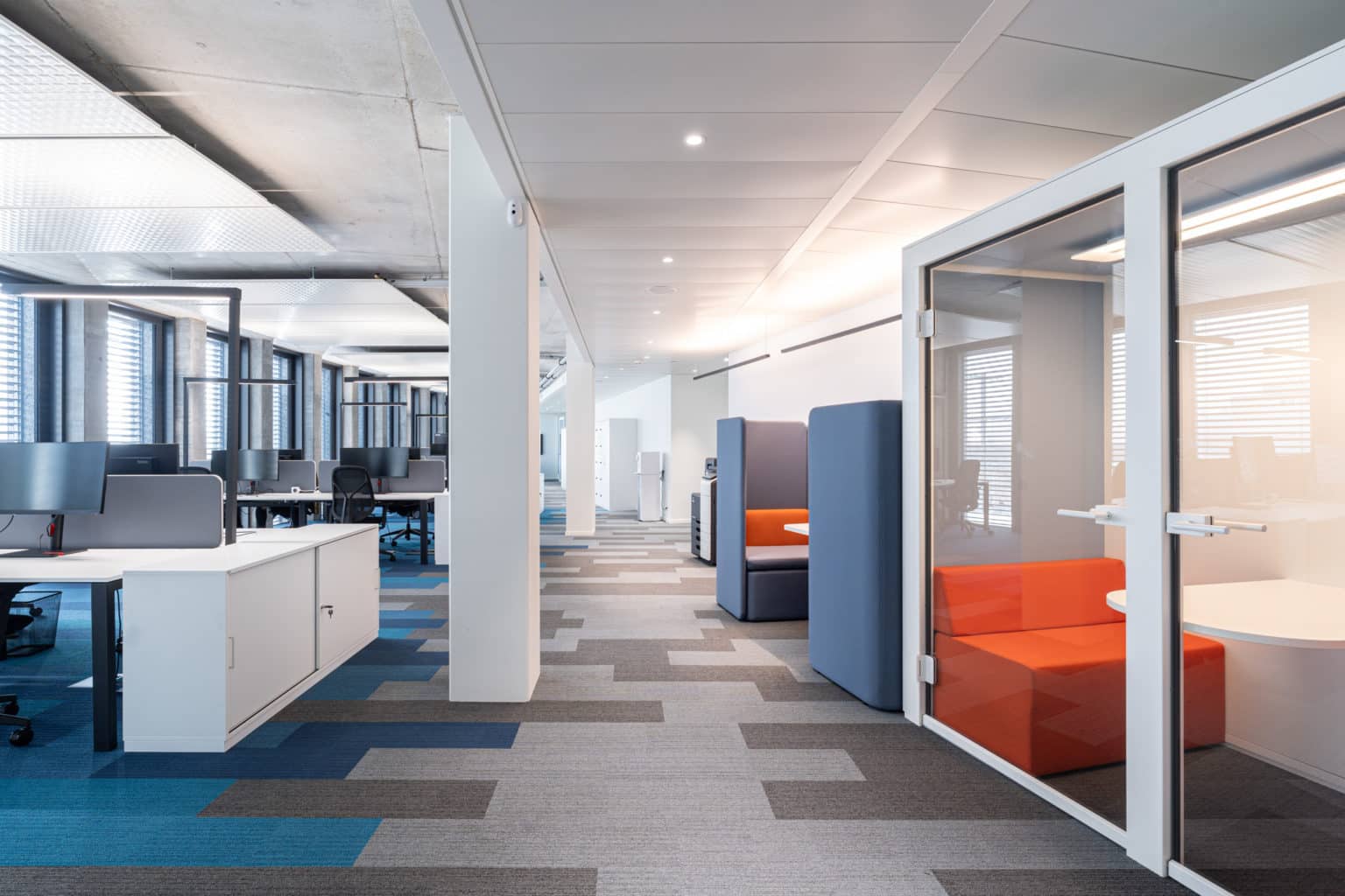 Grand open space moderne doté d'une cabine insonorisée vitrée avec banquette orange et espaces de travail modulaires à Genève.