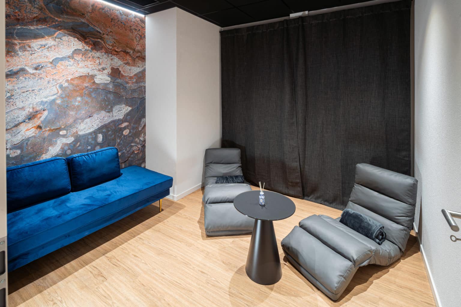 Salon de détente avec canapé en velours bleu, fauteuils lounge, table basse et revêtement mural à l'effet minéral.