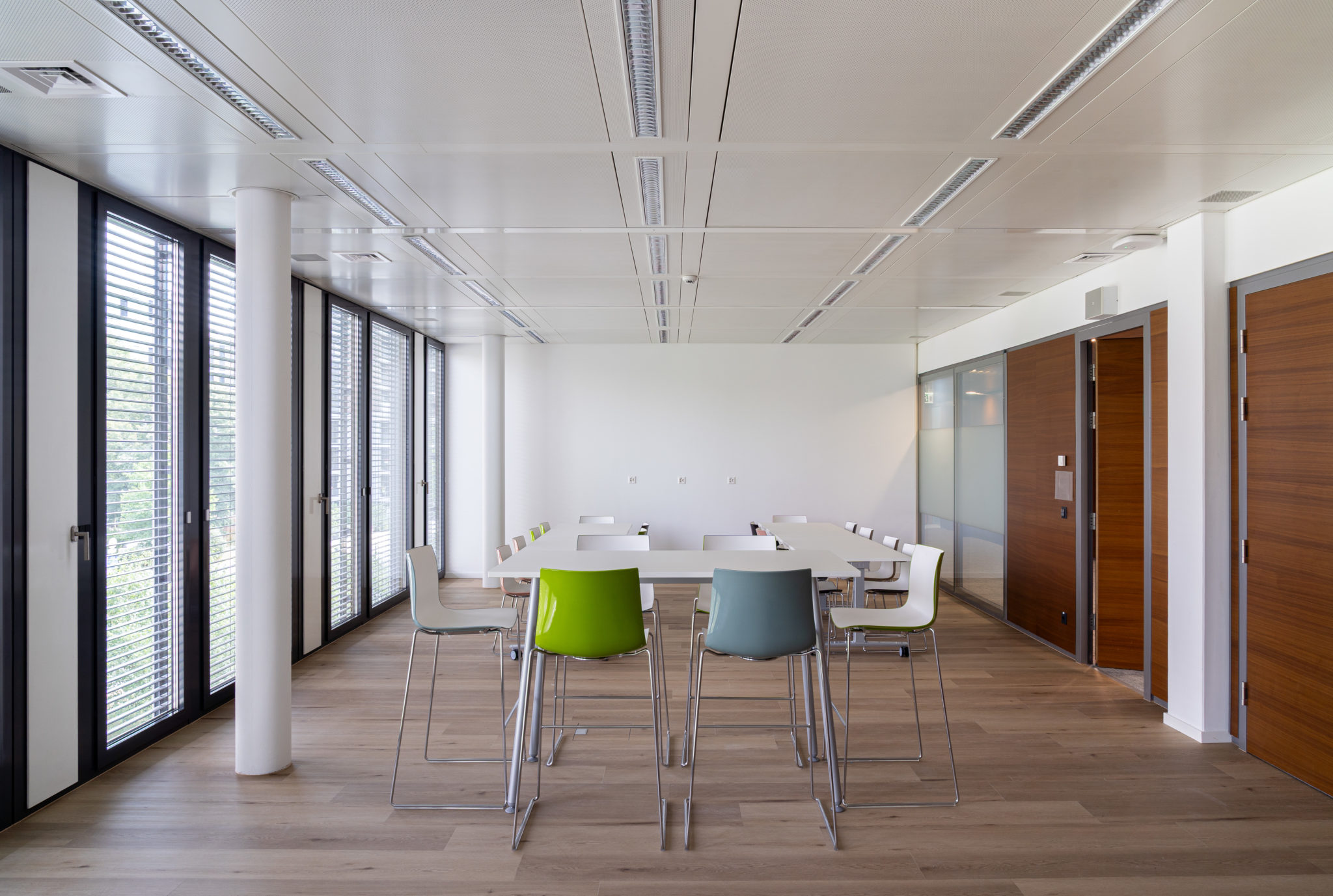 Espace de restauration avec grandes tables hautes, chaises colorées et façade entièrement vitrée.