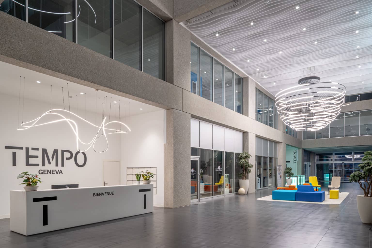 Hall d'accueil contemporain avec banque de réception, espace lounge, suspension lumineuse et circulation centrale.