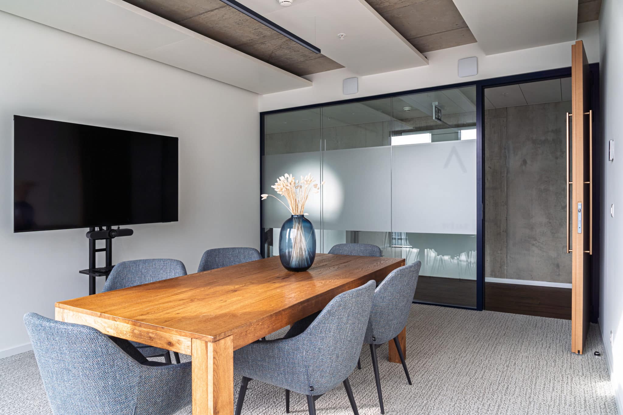 Salle de conférence avec grande table en bois, écran de visioconférence et cloison vitrée.