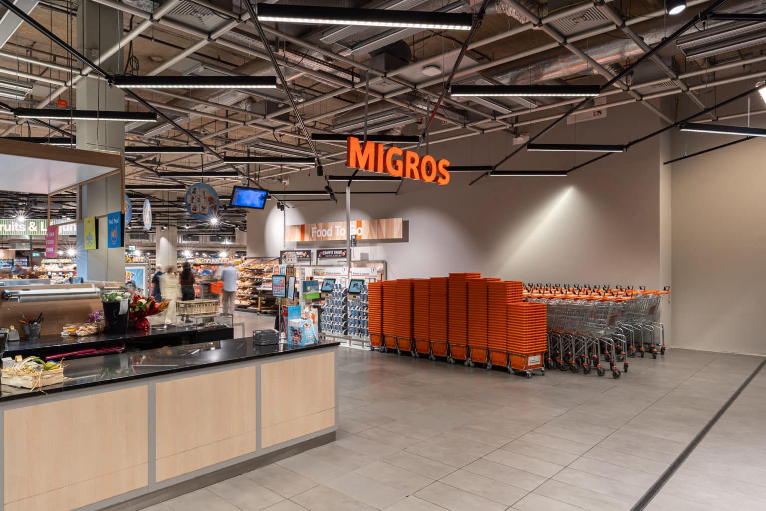 Photographie du supermarché Migros à Nyon, mettant en valeur les espaces d’accueil, le mobilier en bois et l’éclairage intégré.